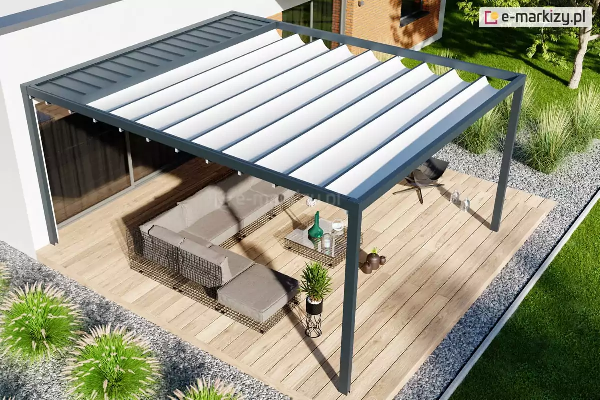 Pergola tarasowa wolnostojąca Luxury, przedstawiona w ujęciu z góry pod kątem. Konstrukcja o grafitowym kolorze posiada ruchome, segmentowe zadaszenie wykonane z białego materiału, które jest rozwinięte. W tylnej części dachu widoczny jest stały panel ochronny z ciemnej blachy, pod którym chowa się poszycie po złożeniu. Pergola zamontowana jest na tarasie z jasnego drewna, na którym ustawiono duży narożnik ogrodowy w kolorze beżowym oraz szklany stolik kawowy. Taras otoczony jest szerokim pasem szarego żwiru dekoracyjnego, w którym symetrycznie posadzono ozdobne kępy traw. W tle widać fragment nowoczesnej bryły budynku o białych ścianach z dużymi oknami oraz fragment zielonego trawnika i drzewa. Perspektywa lotu ptaka dokładnie ukazuje proporcje konstrukcji względem strefy wypoczynkowej oraz estetyczne połączenie materiałów wykończeniowych.
