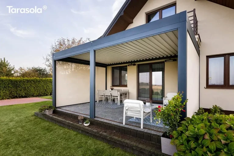 Nowoczesna zadaszona konstrukcja tarasowa Essential Tarasola w kolorze ciemnoantracytowym została zamontowana bezpośrednio do elewacji piętrowego domu jednorodzinnego, tworząc spójną strefę wypoczynkową. Zadaszenie modelu Essential Tarasola posiada płaski dach wykonany z ruchomych, aluminiowych lameli w kolorze białym, które na zdjęciu znajdują się w pozycji całkowicie zamkniętej, co gwarantuje pełną ochronę przed słońcem oraz opadami. Cała konstrukcja opiera się na trzech masywnych, pionowych słupach o kwadratowym przekroju, z których dwa podtrzymują narożniki zewnętrzne, a trzeci umieszczony centralnie dzieli przestrzeń tarasu na dwa równe segmenty. Lewa boczna ściana pergoli została wyposażona w zintegrowaną przesłonę tkaninową typu screen w jasnym kolorze, która pełni funkcję osłony przeciwwiatrowej i wizualnej. Pod pergolą Essential Tarasola zaaranżowano przestronną strefę jadalnianą i relaksacyjną na podłożu z szarej kostki brukowej. W głębi, pod oknem budynku, ustawiono duży biały stół jadalniany z kompletem sześciu nowoczesnych krzeseł o jasnych siedziskach, umieszczonych na dekoracyjnym dywanie zewnętrznym z niebiesko-szarym geometrycznym wzorem. W przedniej części tarasu znajdują się dwa niskie, białe fotele ogrodowe z grubymi, komfortowymi poduszkami, ustawione na drugim, mniejszym dywanie o podobnej stylistyce. Na elewacji budynku, bezpośrednio pod konstrukcją dachu, zamontowano dwa podłużne promienniki ciepła oraz punkty oświetleniowe, co pozwala na korzystanie z tarasu również wieczorami. Taras jest wyniesiony o jeden stopień ponad poziom ogrodu i wykończony ciemną, betonową palisadą o regularnym kształcie. Wokół konstrukcji rozciąga się równo przystrzyżony, zielony trawnik, a po prawej stronie na pierwszym planie widoczne są gęste nasadzenia krzewów oraz donica z niską roślinnością. Bryła budynku charakteryzuje się jasną, beżową elewacją, brązową stolarką okienną oraz ciemnobrązową podbitką dachową.