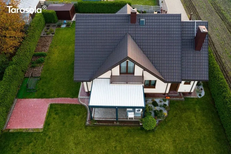 Zdjęcie wykonane z perspektywy lotu ptaka (widok z góry) przedstawia budynek mieszkalny z ciemnobrązowym dachem pokrytym dachówką, do którego przylega nowoczesne zadaszenie tarasu Essential tarasola. Konstrukcja pergoli ma ciemnoantracytowy stelaż i dach wykonany z białych lameli, które są całkowicie zamknięte, tworząc jednolitą jasną płaszczyznę. Taras pod pergolą jest wyłożony jasną kostką brukową i wyposażony w meble ogrodowe: biały stół jadalniany z krzesłami po lewej stronie oraz białą sofę i fotelem po stronie prawej. Teren wokół budynku jest starannie zagospodarowany – bezpośrednio przy tarasie znajduje się rabata z roślinnością ozdobną wysypana jasnym kamieniem, a dalej rozciąga się soczyście zielony trawnik. Po lewej stronie widoczna jest ścieżka z czerwonej kostki brukowej prowadząca do niewielkiego placu z tego samego materiału. Całość posesji jest ogrodzona gęstym, zielonym żywopłotem, a za budynkiem widoczny jest fragment drogi dojazdowej oraz sąsiednie pole uprawne.