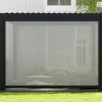 Zdjęcie prezentuje nowoczesną, wolnostojącą pergolę o aluminiowej konstrukcji w kolorze antracytowym, ustawioną na zielonym trawniku w ogrodzie. Frontowa ściana pergoli została w całości przesłonięta pionową roletą zewnętrzną typu screen w kolorze jasnoszarym. Tkanina rolety charakteryzuje się drobnym splotem, który zapewnia skuteczną ochronę przed słońcem i wiatrem, oferując jednocześnie wysoką przezierność – przez opuszczoną osłonę wyraźnie widać wnętrze pergoli z meblami wypoczynkowymi, stołem oraz dekoracyjnymi poduszkami. Dolna krawędź rolety wykończona jest solidną, ciemną listwą obciążającą, która stabilizuje materiał. Zadaszenie konstrukcji jest płaskie, wykonane z ruchomych lameli ustawionych w pozycji lekko uchylonej, z widocznymi nad pergolą fragmentami lameli. W tle widoczny jest fragment białej elewacji budynku z dużymi przeszkleniami oraz zadbana roślinność ogrodowa, w tym krzewy i donica z zieloną rośliną na pierwszym planie.