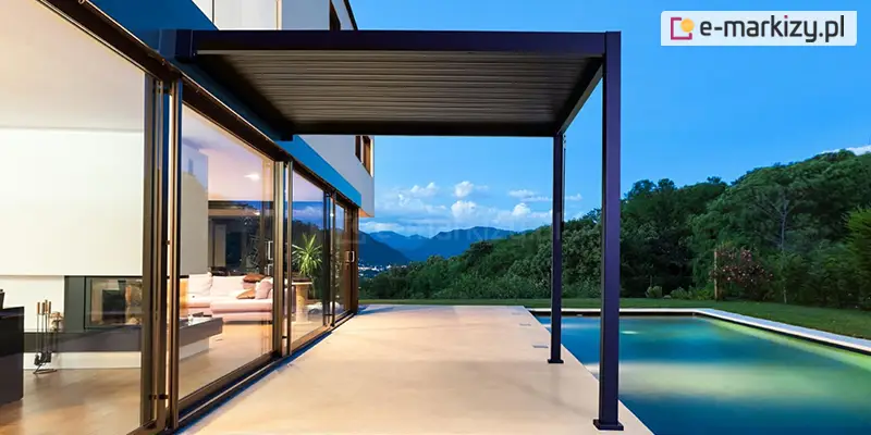 Nowoczesna pergola przyścienna w kolorze antracytowym, zamontowana nad luksusowym tarasem z basenem. Konstrukcja zadaszenia z zamkniętymi lamelami jest przytwierdzona jedną stroną do elewacji piętrowego budynku, a drugą wsparta na dwóch smukłych, kwadratowych słupach. Pod pergolą znajduje się gładka, jasna nawierzchnia tarasu, która sąsiaduje z przeszkloną ścianą salonu, w którym widoczna jest jasna sofa i oświetlone wnętrze. W tle rozpościera się panoramiczny widok na zalesione wzgórza i góry pod wieczornym niebem. Basen z podświetloną, błękitną wodą znajduje się po prawej stronie, bezpośrednio przy krawędzi tarasu. Całość tworzy minimalistyczną i elegancką strefę relaksu o zmierzchu.