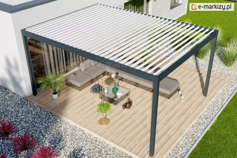 Nowoczesna pergola tarasowa w wersji wolnostojącej, zintegrowana z bryłą białego domu o minimalistycznej architekturze. Konstrukcja o antracytowym stelażu i białym dachu lamelowym osłania duży taras wyłożony deskami kompozytowymi w kolorze naturalnego drewna. Na tarasie znajduje się narożny zestaw wypoczynkowy z szarymi siedziskami, stolik kawowy oraz dekoracyjne rośliny w doniczkach, w tym palma i paproć. Otoczenie tarasu jest starannie zaprojektowane z wykorzystaniem jasnego żwiru, kęp traw ozdobnych oraz bordowych krzewów, a w oddali widać intensywnie zielony trawnik. Widok z góry pozwala na ekspozycję całego układu strefy relaksu oraz płynnego przejścia między wnętrzem domu a ogrodem.