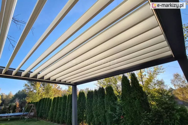 Widok od dołu na nowoczesną pergolę tarasową z systemem ruchomych lameli, zamontowaną w ogrodzie. Konstrukcja wspierająca pergolę, obejmująca widoczne ramy i pionowy słup nośny, wykonana jest z aluminium malowanego proszkowo na kolor ciemnoszary o matowym wykończeniu. Zadaszenie składa się z równolegle ułożonych, szerokich lameli w kolorze białym lub bardzo jasnoszarym. Lamele są w trakcie otwierania, co tworzy efekt stopniowania: po lewej stronie zdjęcia są ustawione pionowo, ukazując czyste, błękitne niebo, natomiast w środkowej i prawej części nachodzą na siebie, tworząc szczelną płaszczyznę dachu. Pod pergolą roztacza się widok na zadbany ogród, w którym wzdłuż granicy posesji posadzono wysoki, gęsty żywopłot z tui. W tle widoczne są liściaste drzewa w jesiennych barwach złota i zieleni oraz fragmenty błękitnego nieba. Po lewej stronie, na trawniku za żywopłotem, dostrzec można elementy wyposażenia rekreacyjnego, w tym drewnianą huśtawkę ogrodową oraz fragment trampoliny. Perspektywa zdjęcia podkreśla geometryczny rytm lameli oraz kontrast między techniczną, surową formą zadaszenia a organiczną roślinnością ogrodu w słoneczny dzień.