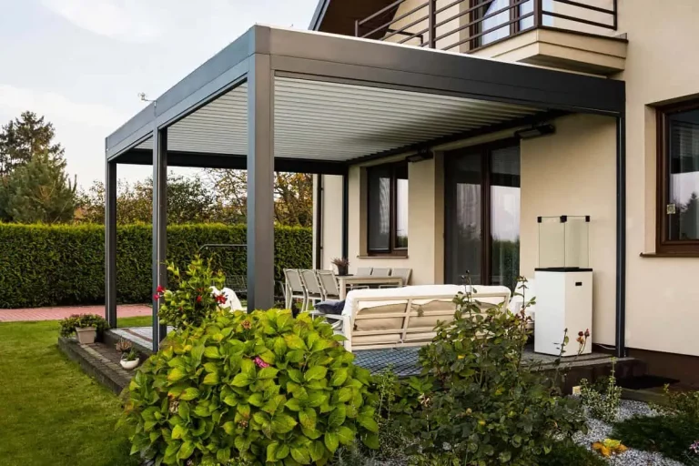 Zdjęcie prezentuje boczne ujęcie nowoczesnej pergoli bioklimatycznej Essential tarasola zamontowanej przy jasnej elewacji domu jednorodzinnego. Konstrukcja w kolorze ciemnego antracytu wspiera dach wykonany z białych, aluminiowych lameli, które są w pełni zamknięte, tworząc szczelną osłonę. Pod zadaszeniem znajduje się zaaranżowana strefa wypoczynkowa z białą sofą ustawioną na niebiesko-białym dywanie zewnętrznym oraz biały stół jadalniany widoczny w głębi. Na tarasie, tuż przy bocznej krawędzi pergoli, stoi wysoki, biały postument zwieńczony szklaną obudową. Pierwszy plan zajmuje bujna roślinność ogrodowa z dużym, zielonym krzewem o różowych kwiatach oraz rabatą wysypaną jasnym żwirem, na której rosną niskie rośliny ozdobne i wrzosy. Całość otacza zadbany, zielony trawnik, a w tle widoczny jest gęsty, wysoki żywopłot oraz fragmenty drzew i sąsiedniej zabudowy pod jasnym, popołudniowym niebem.