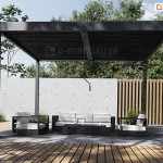 Prosta, minimalistyczna wolnostojąca pergola tarasowa Marlo Duo o konstrukcji z aluminium w kolorze ciemnego antracytu, ustawiona na przestronnym tarasie wyłożonym dużymi, szarymi płytami. Zadaszenie pergoli składa się z dwóch sekcji ruchomych lameli, które są całkowicie zamknięte, tworząc zwartą, płaską powierzchnię chroniącą przed słońcem i deszczem. Pośrodku przedniej belki poprzecznej zamontowana jest pionowa korba manualna służąca do regulacji kąta nachylenia lameli. Pod zadaszeniem zaaranżowano elegancką strefę wypoczynkową z meblami w kontrastowej kolorystyce: czarne ramy sof i foteli zestawiono z grubymi, białymi poduchami siedziskowymi i oparciowymi. W centrum znajduje się niska, czarna ława kawowa o prostokątnym kształcie. W tle widoczne jest nowoczesne ogrodzenie łączące dwa style – po lewej stronie znajduje się ściana z jasnoszarego betonu architektonicznego z charakterystycznymi otworami technologicznymi, a po prawej pionowe, gęsto rozstawione lamele z jasnego drewna. Pomiędzy elementami ogrodzenia oraz przy stopach pergoli rosną ozdobne trawy i niskie krzewy, a nad konstrukcją górują korony liściastych drzew na tle jasnego nieba. Na pierwszym planie widoczny jest fragment podestu z drewnianych desek tarasowych, na który pada cień okolicznych drzew.