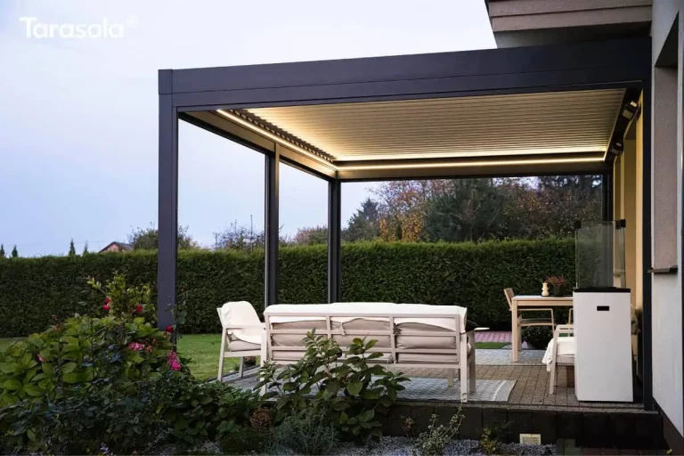 Zdjęcie prezentuje boczną perspektywę tarasu z nowoczesną pergolą bioklimatyczną Essential tarasola o zmierzchu. Konstrukcja o ciemnoantracytowych profilach jest wyposażona w zintegrowane oświetlenie LED w formie liniowych pasów biegnących wzdłuż wewnętrznej ramy, które emituje ciepłe światło, oświetlając spód białych lameli dachowych oraz całą strefę wypoczynkową. Pod zadaszeniem, na jasnej kostce brukowej i wzorzystym dywanie, stoi biała sofa z jasnym obiciem, skierowana tyłem do obiektywu, a w głębi widoczny jest stół jadalniany z krzesłami. Po prawej stronie, przy ścianie budynku, znajduje się wysoki, biały postument z szklaną nadstawką. Po lewej stronie pergoli rośnie rozłożysty krzew z różowymi kwiatami, a tło stanowi wysoki, gęsty żywopłot oddzielający posesję od sąsiednich drzew i zabudowań widocznych pod szarym, wieczornym niebem.