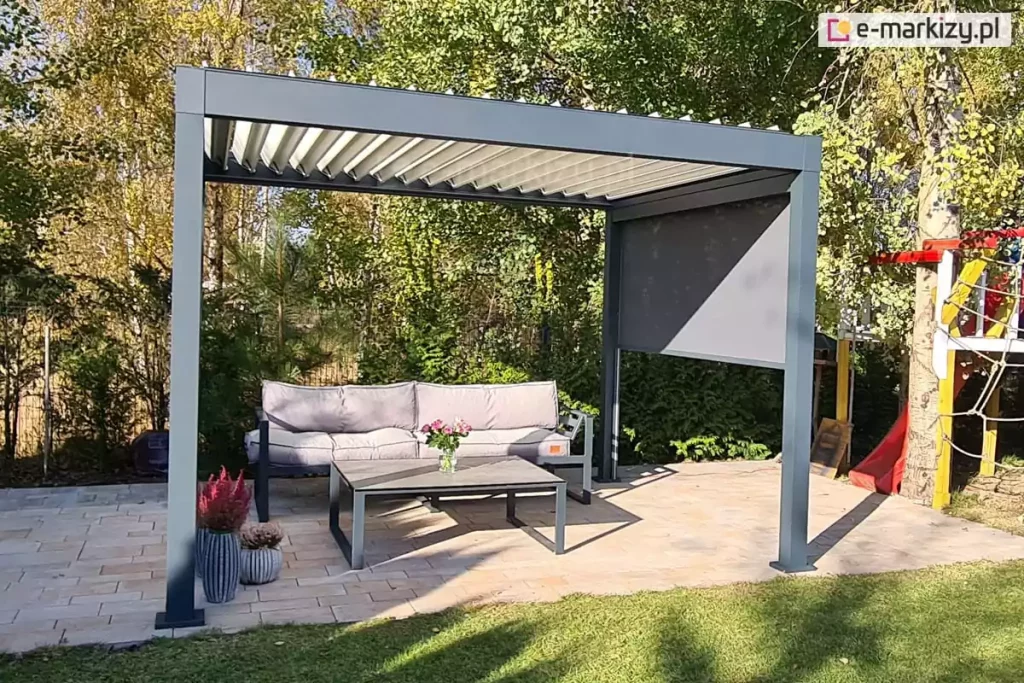Wolnostojąca pergola bioklimatyczna o konstrukcji aluminiowej w kolorze ciemnego antracytu, zaprezentowana w świetle dziennym na przydomowym tarasie. Dach pergoli składa się z ruchomych lameli w kolorze jasnoszarym lub kremowym, które są ustawione pod kątem, ukazując mechanizm otwierania zadaszenia. Na jednej z bocznych ścian zamontowano roletę zewnętrzną typu screen w kolorze grafitowym, która jest rozwinięta do połowy wysokości, rzucając wyraźny, geometryczny cień na wnętrze konstrukcji. Pod pergolą, na podłożu z jasnej kostki brukowej o beżowo-piaskowej barwie, urządzono strefę relaksu z sofą ogrodową wyposażoną w szare poduszki oraz stolikiem kawowym z ciemnym stelażem, na którym stoi wazon z różowymi kwiatami. W lewym dolnym rogu widoczne są ozdobne donice z wrzosami. Tło stanowi gęsta roślinność ogrodowa z drzewami o zielono-żółtych liściach oraz fragment kolorowego placu zabaw z czerwoną zjeżdżalnią. Całość kompozycji podkreśla funkcjonalność pergoli jako systemu ochrony przed słońcem w ogrodzie.