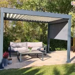 Wolnostojąca pergola bioklimatyczna o konstrukcji aluminiowej w kolorze ciemnego antracytu, zaprezentowana w świetle dziennym na przydomowym tarasie. Dach pergoli składa się z ruchomych lameli w kolorze jasnoszarym lub kremowym, które są ustawione pod kątem, ukazując mechanizm otwierania zadaszenia. Na jednej z bocznych ścian zamontowano roletę zewnętrzną typu screen w kolorze grafitowym, która jest rozwinięta do połowy wysokości, rzucając wyraźny, geometryczny cień na wnętrze konstrukcji. Pod pergolą, na podłożu z jasnej kostki brukowej o beżowo-piaskowej barwie, urządzono strefę relaksu z sofą ogrodową wyposażoną w szare poduszki oraz stolikiem kawowym z ciemnym stelażem, na którym stoi wazon z różowymi kwiatami. W lewym dolnym rogu widoczne są ozdobne donice z wrzosami. Tło stanowi gęsta roślinność ogrodowa z drzewami o zielono-żółtych liściach oraz fragment kolorowego placu zabaw z czerwoną zjeżdżalnią. Całość kompozycji podkreśla funkcjonalność pergoli jako systemu ochrony przed słońcem w ogrodzie.