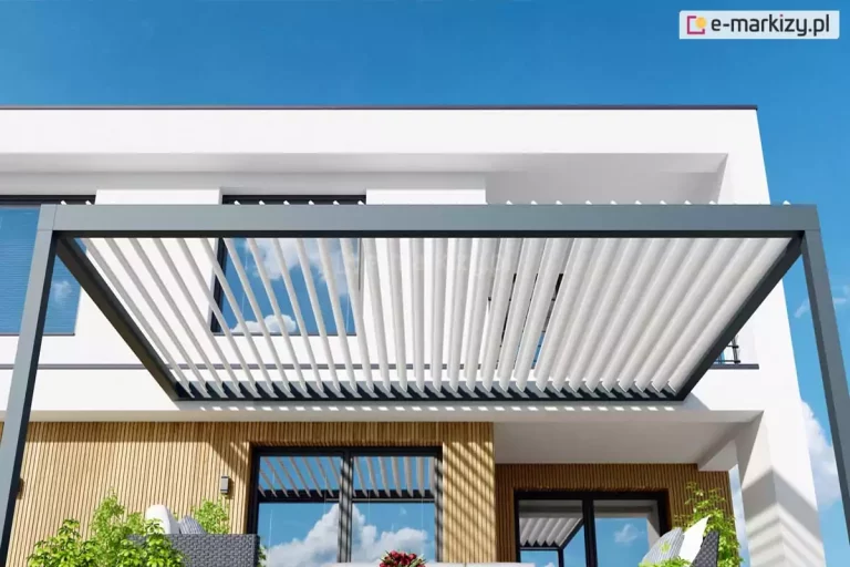 Nowoczesna pergola tarasowa o konstrukcji aluminiowej zamontowana przy elewacji piętrowego budynku o białej fasadzie. Konstrukcja pergoli składa się z ciemnoszarej, niemal czarnej ramy wspartej na pionowych słupach o kwadratowym przekroju. Zadaszenie tworzy system ruchomych, obrotowych lameli w kolorze białym, które w tej konfiguracji są częściowo otwarte, ustawione pod kątem, co pozwala na przenikanie promieni słonecznych i ukazuje błękitne niebo z delikatnymi chmurami. Ciemna rama pergoli kontrastuje z jasnym kolorem lameli oraz białą ścianą budynku. Poniżej zadaszenia widoczna jest elewacja parteru wykończona pionowymi lamelami dekoracyjnymi w kolorze naturalnego drewna. W ścianie osadzone są duże, ciemne okna tarasowe, w których odbija się niebo oraz konstrukcja pergoli. Na tarasie znajdują się meble ogrodowe z ciemnej plecionki z szarymi poduszkami oraz zielona roślinność w donicach. Całość przedstawiona jest w pełnym świetle dziennym przy bezchmurnym, intensywnie niebieskim niebie, co podkreśla nowoczesny design i funkcjonalność przydomowej strefy wypoczynku.