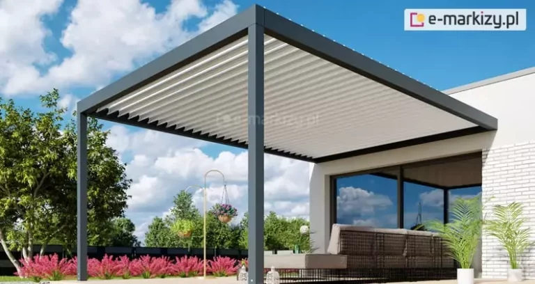 Nowoczesna pergola lamelowa przyścienna BIoline zamontowana przy elewacji budynku o estetyce białej cegły. Konstrukcja pergoli wykonana jest z profili aluminiowych w kolorze ciemnego antracytu i opiera się na dwóch pionowych słupach nośnych o kwadratowym przekroju. Zadaszenie bioklimatyczne składa się z białych lameli, które są ukazane w pozycji lekko uchylonej, demonstrując mechanizm regulacji osłony przeciwsłonecznej. Pod pergolą znajduje się strefa wypoczynkowa z szarymi, modułowymi sofami o ażurowej konstrukcji, a obok nich ustawiono białe donice z roślinami egzotycznymi, takimi jak palmy. W tle widoczny jest ogród z pasem różowych krzewów ozdobnych, zielone drzewa liściaste oraz czarne, nowoczesne ogrodzenie panelowe. Przeszklone, duże drzwi tarasowe odbijają błękitne niebo z białymi chmurami, tworząc spójną całość z otoczeniem zewnętrznym. Perspektywa zdjęcia z dołu podkreśla wysokość i solidność aluminiowej konstrukcji zadaszenia.