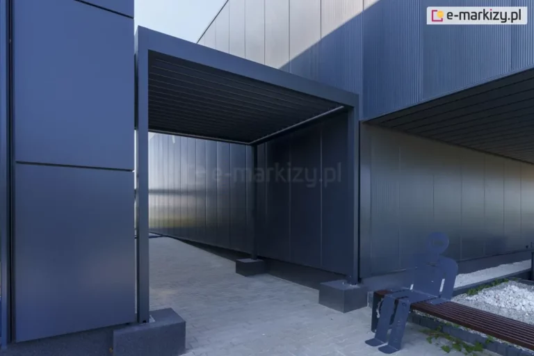 Przejście między budynkami osłonięte antracytową pergolą lamelową SB400 Selt z zamkniętym zadaszeniem z piór aluminiowych. W pobliżu przejścia ustawiono ławeczkę drewnianą z siedzącym na niej płaskim ludzikiem z aluminium w kolorze antracytowym.