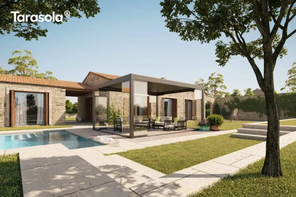 Nowoczesna pergola lamelowa essential Tarasola w ciemnym, antracytowym kolorze, stanowiąca wolnostojący element zabudowy tarasu wykonanego z jasnych płyt kamiennych. Konstrukcja wyposażona jest w dach z ruchomymi lamelami oraz szklane systemy przesuwne, które domykają boczne ściany, tworząc luksusowy ogród zimowy. Wewnątrz pergoli, na podłożu z drobnego żwiru, ustawiony jest komplet wypoczynkowy składający się z ciemnych foteli i sofy z jasnymi poduszkami oraz stolika kawowego. Instalacja znajduje się w sąsiedztwie basenu o błękitnej wodzie i przylega wizualnie do parterowego budynku z elewacją z naturalnego kamienia, ozdobionego drewnianymi okiennicami. Całość otacza zadbana zieleń ogrodowa, trawnik, kamienne schody oraz drzewa liściaste, a w tle widoczny jest wysoki mur z kamienia, co tworzy spójną i elegancką strefę rekreacyjną pod bezchmurnym niebem.