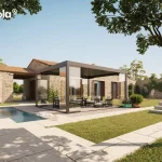 Nowoczesna pergola lamelowa essential Tarasola w ciemnym, antracytowym kolorze, stanowiąca wolnostojący element zabudowy tarasu wykonanego z jasnych płyt kamiennych. Konstrukcja wyposażona jest w dach z ruchomymi lamelami oraz szklane systemy przesuwne, które domykają boczne ściany, tworząc luksusowy ogród zimowy. Wewnątrz pergoli, na podłożu z drobnego żwiru, ustawiony jest komplet wypoczynkowy składający się z ciemnych foteli i sofy z jasnymi poduszkami oraz stolika kawowego. Instalacja znajduje się w sąsiedztwie basenu o błękitnej wodzie i przylega wizualnie do parterowego budynku z elewacją z naturalnego kamienia, ozdobionego drewnianymi okiennicami. Całość otacza zadbana zieleń ogrodowa, trawnik, kamienne schody oraz drzewa liściaste, a w tle widoczny jest wysoki mur z kamienia, co tworzy spójną i elegancką strefę rekreacyjną pod bezchmurnym niebem.