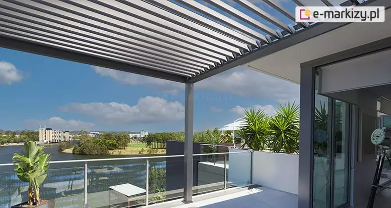 Nowoczesna pergola lamelowa wolnostojąca SB350 Aluprof zainstalowana na przestronnym tarasie widokowym. Zdjęcie prezentuje antracytową konstrukcję z dachem wykonanym z ruchomych, jasnoszarych lameli ustawionych pod kątem. Pergola przylega do elewacji budynku i jest wsparta na pionowym słupie nośnym. Taras zabezpieczony jest szklaną balustradą, zza której rozpościera się widok na rzekę, zielone nabrzeże oraz zabudowę miejską pod błękitnym niebem z białymi chmurami. Na tarasie widoczne są rośliny egzotyczne w donicach, w tym juka i strelicja, co podkreśla luksusowy charakter strefy wypoczynku.