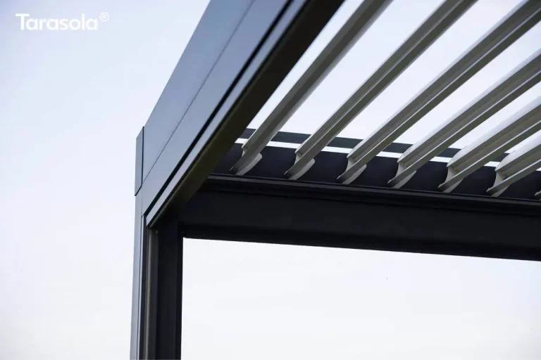 Zdjęcie przedstawia zbliżenie na górny narożnik i detale techniczne konstrukcji dachu modelu Essential tarasola. Na pierwszym planie widoczne jest solidne łączenie pionowego słupa z poziomą belką ramy, wykonane z aluminium malowanego proszkowo na kolor ciemnego antracytu o matowym wykończeniu. Powyżej ramy zamontowany jest system zadaszenia bioklimatycznego składający się z szeregu ruchomych, białych lameli o opływowym kształcie. Lamele są w tym momencie uchylone pod kątem, co ukazuje mechanizm ich osadzenia w bocznych profilach prowadzących. Do pionowej podpory zamocowana została prowadnica refleksola Zip. Całość detalu układa się na tle jasnego, niemal białego nieba, co podkreśla czyste linie i nowoczesny, minimalistyczny design aluminiowej pergoli.