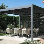 Nowoczesna wolnostojąca pergola ogrodowa Marlo Duo o aluminiowej konstrukcji w kolorze ciemnego antracytu, ustawiona na jasnym, betonowym patio w otoczeniu gęstej zieleni. Zadaszenie pergoli składa się z poziomych lameli, które są lekko otwarte, tworząc osłonę przed słońcem oraz wyraźny cień na podłodze tarasu. Konstrukcja wsparta jest na czterech solidnych kwadratowych słupach, a jedna z bocznych ścian została zabudowana przesuwnymi panelami typu shutters z poziomymi lamelami w tym samym kolorze co stelaż. Wewnątrz pergoli znajduje się jadalniana strefa ogrodowa z dużym stołem o drewnianym blacie, przy którym ustawiono eleganckie fotele z zaokrąglonymi oparciami wykonanymi z jasnoszarego sznurka technorattanowego. Na stole widoczna jest dekoracyjna zastawa oraz wazon z kwiatami. Tło zdjęcia stanowi wysoki, starannie przystrzyżony żywopłot oraz korony drzew pod czystym, błękitnym niebem. Po prawej stronie widoczny jest fragment ścieżki wykonanej z czerwonej kostki brukowej oraz niska roślinność ogrodowa. Całość kompozycji podkreśla luksusowy i nowoczesny charakter przydomowej strefy wypoczynku.