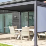 Fotografia ukazuje nowoczesną pergolę tarasową w kolorze antracytowym, zamontowaną przy budynku o ciemnej elewacji i dużych przeszkleniach. Aluminiowa konstrukcja wyposażona jest w poziomą ramę zadaszenia oraz pionową osłonę boczną w formie rolety typu screen w kolorze jasnoszarym. Roleta jest częściowo opuszczona, zasłaniając górną połowę bocznego światła pergoli, co zapewnia ochronę przed słońcem przy zachowaniu widoczności. Na tarasie wyłożonym jasnym drewnem ustawiono biały stół jadalniany oraz sześć krzeseł z jasnej plecionki z miękkimi siedziskami. Całość aranżacji tworzy spójną, minimalistyczną strefę jadalnianą na świeżym powietrzu, sąsiadującą z zadbanym trawnikiem.