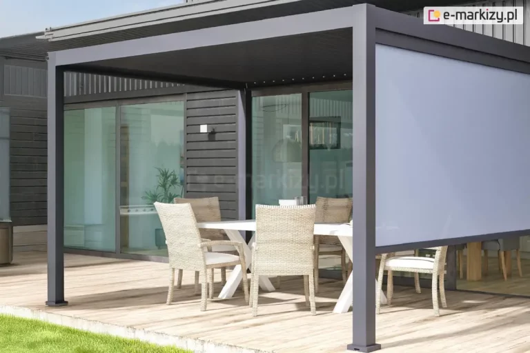 Fotografia ukazuje nowoczesną pergolę tarasową w kolorze antracytowym, zamontowaną przy budynku o ciemnej elewacji i dużych przeszkleniach. Aluminiowa konstrukcja wyposażona jest w poziomą ramę zadaszenia oraz pionową osłonę boczną w formie rolety typu screen w kolorze jasnoszarym. Roleta jest częściowo opuszczona, zasłaniając górną połowę bocznego światła pergoli, co zapewnia ochronę przed słońcem przy zachowaniu widoczności. Na tarasie wyłożonym jasnym drewnem ustawiono biały stół jadalniany oraz sześć krzeseł z jasnej plecionki z miękkimi siedziskami. Całość aranżacji tworzy spójną, minimalistyczną strefę jadalnianą na świeżym powietrzu, sąsiadującą z zadbanym trawnikiem.