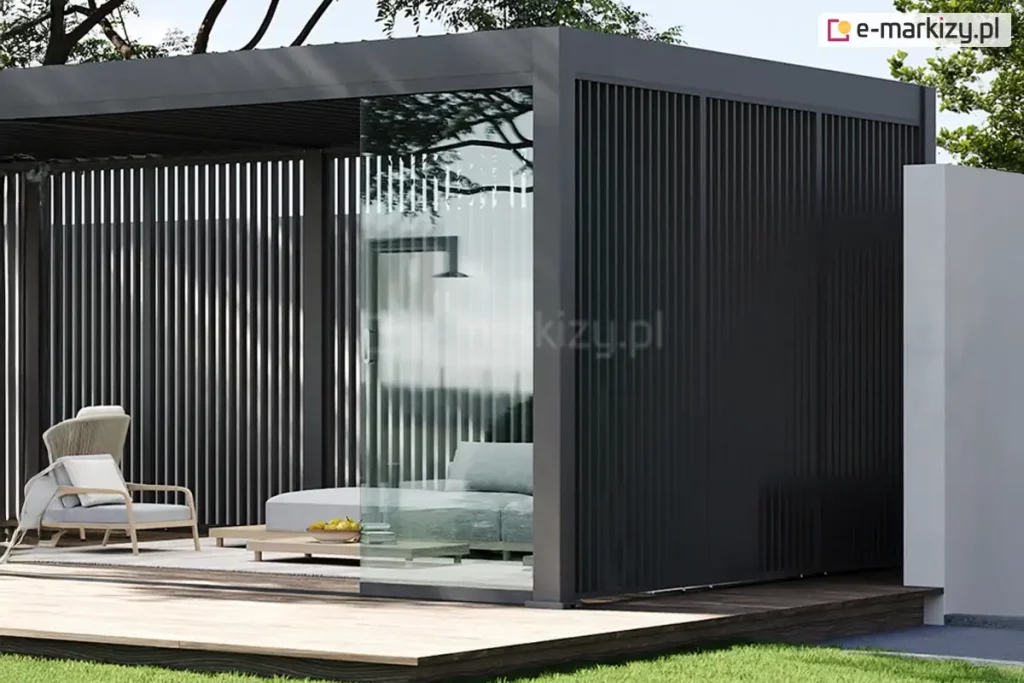 Zdjęcie ukazuje rozbudowaną konfigurację pergoli Marlo w kolorze ciemnego antracytu, posadowioną na ciemnym drewnianym tarasie. Konstrukcja charakteryzuje się pełną zabudową boczną wykonaną z gęstych, pionowych profili aluminiowych, które tworzą efekt żaluzji stałej. Frontowa część pergoli posiada przesuwne systemy szklenia, z których jeden panel jest częściowo otwarty, ukazując wnętrze wyposażone w niskie meble lounge, szare poduszki oraz dekoracyjny stolik z owocami. W tle widoczne są korony drzew oraz fragment białej ściany, co nadaje całości luksusowy i intymny charakter.
