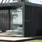 Zdjęcie ukazuje rozbudowaną konfigurację pergoli Marlo w kolorze ciemnego antracytu, posadowioną na ciemnym drewnianym tarasie. Konstrukcja charakteryzuje się pełną zabudową boczną wykonaną z gęstych, pionowych profili aluminiowych, które tworzą efekt żaluzji stałej. Frontowa część pergoli posiada przesuwne systemy szklenia, z których jeden panel jest częściowo otwarty, ukazując wnętrze wyposażone w niskie meble lounge, szare poduszki oraz dekoracyjny stolik z owocami. W tle widoczne są korony drzew oraz fragment białej ściany, co nadaje całości luksusowy i intymny charakter.