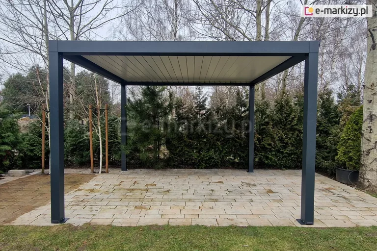 Widok od boku na wolnostojącą pergolę bioklimatyczną Sb350 Selt o antracytowej konstrukcji aluminiowej, ustawioną na tarasie z jasnej kostki brukowej. Pergola posiada cztery kwadratowe słupy nośne oraz dach z zamkniętymi lamelami w kolorze białym, tworzącymi szczelne zadaszenie. Konstrukcja jest zamontowana w ogrodzie; bezpośrednio za nią znajduje się gęsty pas zieleni składający się z wysokich tui, sosen oraz młodych brzóz wspartych na tyczkach. Po prawej stronie widoczny jest pień dużego drzewa liściastego oraz fragment czarnej donicy. Zdjęcie prezentuje symetryczną bryłę pergoli na tle naturalnej roślinności w pochmurny dzień.