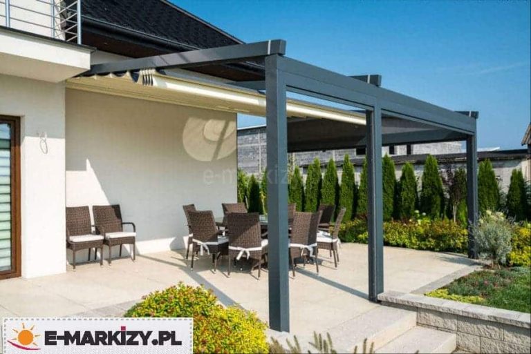 Nowoczesna przyścienna pergola tarasowa o konstrukcji aluminiowej w kolorze ciemnego antracytu, zamontowana bezpośrednio do elewacji białego budynku jednorodzinnego. Zadaszenie składa się z dwóch modułów z jasną, kremową tkaniną rzymską, która na zdjęciu jest częściowo zwinięta przy ścianie budynku. Konstrukcja wsparta jest na trzech pionowych słupach o kwadratowym przekroju, a w jednym z bocznych modułów zainstalowano dodatkową pionową roletę typu screen w ciemnym kolorze, zapewniającą ochronę przed słońcem i wiatrem. Pod pergolą na jasnym, wyłożonym płytami tarasie znajduje się zestaw mebli ogrodowych wykonanych z ciemnobrązowego technorattanu, obejmujący duży stół oraz krzesła z białymi poduszkami siedziskowymi. Taras graniczy z ogrodem, w którym widoczne są niskie krzewy ozdobne oraz wysoki żywopłot z tui oddzielający posesję od sąsiedniego budynku w stanie surowym. Zdjęcie wykonane w słoneczny dzień przy bezchmurnym niebie, prezentuje praktyczne zastosowanie modułowej pergoli jako przedłużenia strefy wypoczynkowej domu.