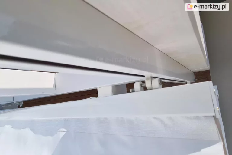 Zbliżenie białej prowadnicy dachu Supro z białym wózkiem 3cm zamocowanym do białek belki przeciwwietrznej z tkaniną Opatex w kolorze białym. Prowadnica zamocowana została do pomalowanej na biało konstrukcji pergoli drewnianej.