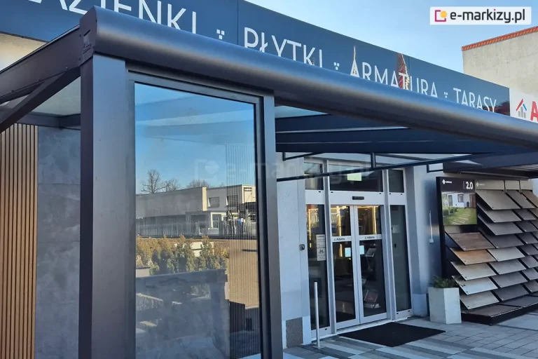 Ekspozycja zewnętrzna nowoczesnej pergoli tarasowej o konstrukcji aluminiowej w kolorze ciemnografitowym. Kluczowym elementem widocznym na pierwszym planie jest stałe przeszklenie boczne, oprawione w gruby profil pionowy, które pełni funkcję osłony przed wiatrem lub ścianki działowej. Nad ścianką stałą widoczna jest rynna zadaszenia o zaokrąglonym kształcie, a po lewej stronie widoczny jest fragment ściany wykończonej pionowymi lamelami drewnianymi oraz szarym spiekiem kwarcowym. W tle znajduje się wejście do salonu sprzedaży z przeszklonymi drzwiami automatycznymi oraz ekspozytor z próbkami materiałów elewacyjnych lub tarasowych. Całość ukazana jest w świetle dziennym, z wyraźnym odbiciem otoczenia i nieba w szybie stałego okna.