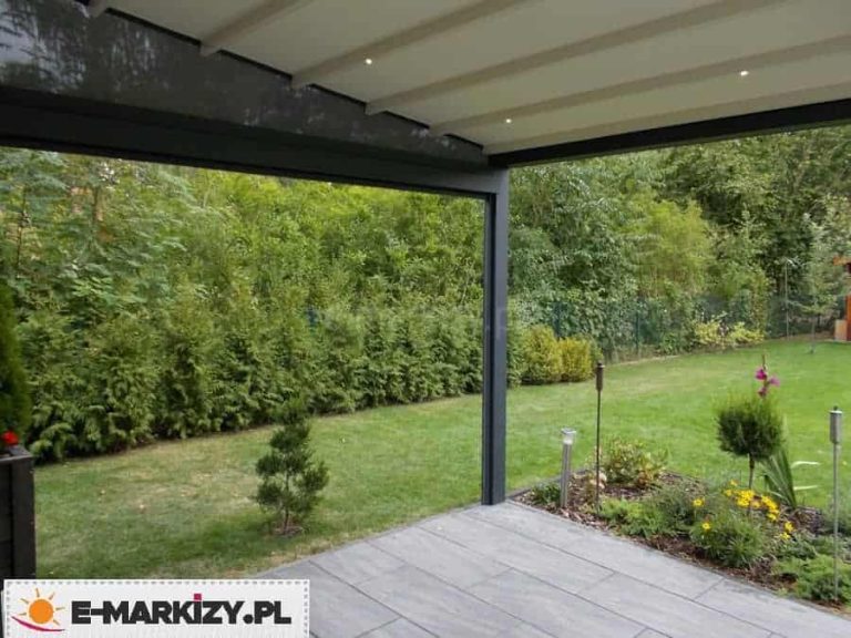 Zdjęcie wykonane z perspektywy osoby znajdującej się pod zadaszeniem pergoli rzymskiej Moderno, ukazujące widok na ogród. Widoczna jest dolna część białego poszycia tkaninowego, w którego profilach zamontowano okrągłe oprawy oświetlenia LED. Konstrukcja wsporcza w kolorze antracytowym obramowuje widok na zielony trawnik oraz gęsty żywopłot z tui i innych drzew liściastych. Taras wyłożony jest szarymi płytami o teksturze imitującej kamień, a bezpośrednio przy jego krawędzi znajduje się rabata z niskimi roślinami, kwiatami oraz ozdobnymi lampami solarnymi w kształcie słupków. Perspektywa podkreśla funkcjonalność oświetlenia zintegrowanego z pergolą, które pozwala na korzystanie z tarasu po zmroku.