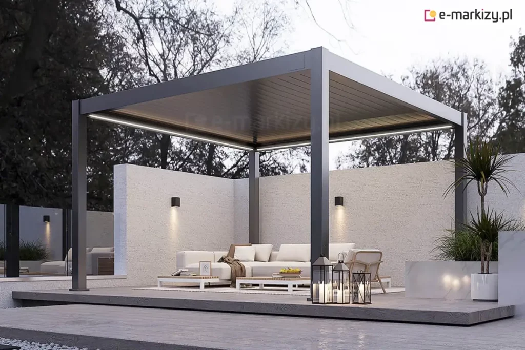 Nowoczesna, wolnostojąca pergola bioklimatyczna Rubio o wymiarach 4x4 metry, wykonana z aluminium w kolorze antracytowym, zintegrowana z minimalistyczną strefą wypoczynku. Konstrukcja z dachem lamelowym w pozycji zamkniętej posiada wbudowane oświetlenie liniowe LED o zimnej barwie, biegnące wzdłuż wewnętrznych krawędzi ramy. Pod pergolą znajduje się luksusowy zestaw mebli ogrodowych: jasna sofa narożna, fotele oraz niskie stoliki kawowe ustawione na jasnym dywanie. Taras został zaprojektowany na podwyższonym podeście z ciemnoszarego kompozytu, otoczonym opaską z szarego grysu dekoracyjnego. Tło stanowi wysoki, biały mur o strukturalnym tynku z zamontowanymi kinkietami elewacyjnymi typu góra-dół oraz gęsta ściana drzew liściastych. W aranżacji wykorzystano roślinność w donicach (m.in. juka) oraz ozdobne lampiony ze świecami umieszczone na krawędzi podestu. Zdjęcie wykonane w porze zmierzchu, co uwypukla kontrast między ciemną bryłą pergoli a systemem jej podświetlenia.