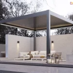 Nowoczesna, wolnostojąca pergola bioklimatyczna Rubio o wymiarach 4x4 metry, wykonana z aluminium w kolorze antracytowym, zintegrowana z minimalistyczną strefą wypoczynku. Konstrukcja z dachem lamelowym w pozycji zamkniętej posiada wbudowane oświetlenie liniowe LED o zimnej barwie, biegnące wzdłuż wewnętrznych krawędzi ramy. Pod pergolą znajduje się luksusowy zestaw mebli ogrodowych: jasna sofa narożna, fotele oraz niskie stoliki kawowe ustawione na jasnym dywanie. Taras został zaprojektowany na podwyższonym podeście z ciemnoszarego kompozytu, otoczonym opaską z szarego grysu dekoracyjnego. Tło stanowi wysoki, biały mur o strukturalnym tynku z zamontowanymi kinkietami elewacyjnymi typu góra-dół oraz gęsta ściana drzew liściastych. W aranżacji wykorzystano roślinność w donicach (m.in. juka) oraz ozdobne lampiony ze świecami umieszczone na krawędzi podestu. Zdjęcie wykonane w porze zmierzchu, co uwypukla kontrast między ciemną bryłą pergoli a systemem jej podświetlenia.