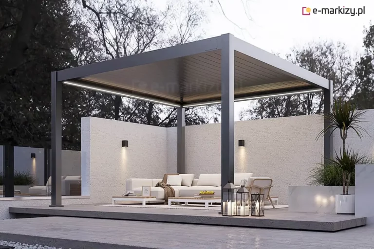 Nowoczesna, wolnostojąca pergola bioklimatyczna Rubio o wymiarach 4x4 metry, wykonana z aluminium w kolorze antracytowym, zintegrowana z minimalistyczną strefą wypoczynku. Konstrukcja z dachem lamelowym w pozycji zamkniętej posiada wbudowane oświetlenie liniowe LED o zimnej barwie, biegnące wzdłuż wewnętrznych krawędzi ramy. Pod pergolą znajduje się luksusowy zestaw mebli ogrodowych: jasna sofa narożna, fotele oraz niskie stoliki kawowe ustawione na jasnym dywanie. Taras został zaprojektowany na podwyższonym podeście z ciemnoszarego kompozytu, otoczonym opaską z szarego grysu dekoracyjnego. Tło stanowi wysoki, biały mur o strukturalnym tynku z zamontowanymi kinkietami elewacyjnymi typu góra-dół oraz gęsta ściana drzew liściastych. W aranżacji wykorzystano roślinność w donicach (m.in. juka) oraz ozdobne lampiony ze świecami umieszczone na krawędzi podestu. Zdjęcie wykonane w porze zmierzchu, co uwypukla kontrast między ciemną bryłą pergoli a systemem jej podświetlenia.