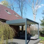 Zdjęcie prezentuje nowoczesną pergolę tarasową Tarasola Flat o konstrukcji aluminiowej w kolorze antracytowym, zamontowaną do elewacji piętrowego budynku mieszkalnego. Zadaszenie stanowi jasny, kremowy dach tkaninowy, który został wyposażony w zintegrowany system oświetlenia punktowego LED, rozmieszczony równomiernie na profilach poprzecznych. Konstrukcja wspiera się na solidnych słupach o kwadratowym przekroju, a pod nią znajduje się przestronny taras wyłożony jasnymi płytami. Na obrzeżach tarasu ustawiono masywne, geometryczne donice wykonane ze stali corten o charakterystycznej, rdzawej barwie, w których posadzono trawę ozdobną oraz formowane krzewy. Budynek charakteryzuje się nowoczesną architekturą z białym tynkiem, dużymi przeszkleniami oraz szklanymi balustradami na balkonach. W tle widoczna jest gęsta roślinność ogrodowa, co podkreśla funkcjonalność systemu w aranżowaniu przydomowych stref wypoczynkowych.