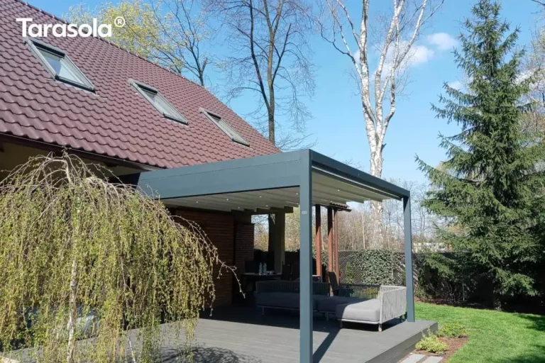 Zdjęcie prezentuje nowoczesną pergolę tarasową Tarasola Flat o konstrukcji aluminiowej w kolorze antracytowym, zamontowaną do elewacji piętrowego budynku mieszkalnego. Zadaszenie stanowi jasny, kremowy dach tkaninowy, który został wyposażony w zintegrowany system oświetlenia punktowego LED, rozmieszczony równomiernie na profilach poprzecznych. Konstrukcja wspiera się na solidnych słupach o kwadratowym przekroju, a pod nią znajduje się przestronny taras wyłożony jasnymi płytami. Na obrzeżach tarasu ustawiono masywne, geometryczne donice wykonane ze stali corten o charakterystycznej, rdzawej barwie, w których posadzono trawę ozdobną oraz formowane krzewy. Budynek charakteryzuje się nowoczesną architekturą z białym tynkiem, dużymi przeszkleniami oraz szklanymi balustradami na balkonach. W tle widoczna jest gęsta roślinność ogrodowa, co podkreśla funkcjonalność systemu w aranżowaniu przydomowych stref wypoczynkowych.