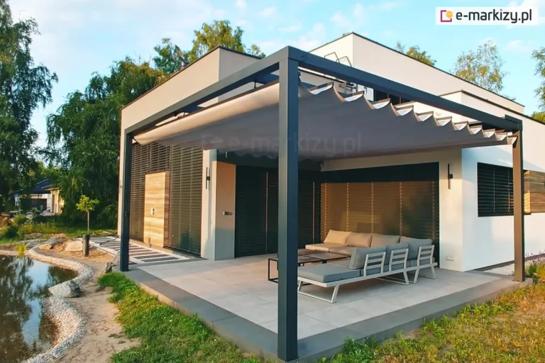 Parterowy domek wypoczynkowy w lesie, tuż przy oczku wodnym o brzegu wyłożonym kamykami, z niewielkim tarasem wyłożonym betonowymi płytami. Do narożników tarasu przymocowano podpory pergoli TAKO z dachem rzymskim rozwiniętym nad narożnikiem z szarymi poduchami i niewielkim stolikiem z jasnym blatem. Ściany domku oświetla zachodzące słońce. W tle widoczne drzewa i sąsiedni domek.
