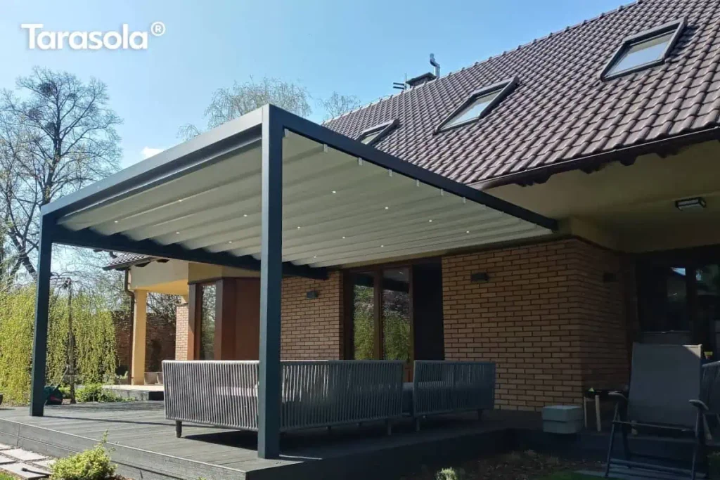 Zdjęcie prezentuje nowoczesną pergolę ogrodową o wymiarach 3x6m, wykonaną z aluminiowej konstrukcji w kolorze antracytowym. Jest to model przyścienny Tarasola Flat, zamontowany do elewacji budynku z jasnobrązowej cegły, przykrytego spadzistym dachem z ciemnej dachówki. Zadaszenie pergoli stanowi rozsuwany dach tkaninowy w kolorze jasnokremowym, który jest całkowicie rozwinięty i wyposażony w zintegrowane, punktowe oświetlenie LED rozmieszczone w regularnych odstępach na profilach poprzecznych. Pod pergolą, na tarasie z ciemnych desek kompozytowych, znajduje się strefa wypoczynkowa z nowoczesną sofą ogrodową o ażurowej konstrukcji z szarego sznurka lub technorattanu. W tle widoczny jest słoneczny ogród z gęstą roślinnością, drzewami liściastymi oraz fragmentem trawnika pod bezchmurnym, błękitnym niebem. Konstrukcja pergoli charakteryzuje się prostymi liniami i solidnymi słupami wsporczymi, tworząc funkcjonalną i estetyczną przestrzeń do relaksu na świeżym powietrzu.