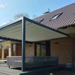 Zdjęcie prezentuje nowoczesną pergolę ogrodową o wymiarach 3x6m, wykonaną z aluminiowej konstrukcji w kolorze antracytowym. Jest to model przyścienny Tarasola Flat, zamontowany do elewacji budynku z jasnobrązowej cegły, przykrytego spadzistym dachem z ciemnej dachówki. Zadaszenie pergoli stanowi rozsuwany dach tkaninowy w kolorze jasnokremowym, który jest całkowicie rozwinięty i wyposażony w zintegrowane, punktowe oświetlenie LED rozmieszczone w regularnych odstępach na profilach poprzecznych. Pod pergolą, na tarasie z ciemnych desek kompozytowych, znajduje się strefa wypoczynkowa z nowoczesną sofą ogrodową o ażurowej konstrukcji z szarego sznurka lub technorattanu. W tle widoczny jest słoneczny ogród z gęstą roślinnością, drzewami liściastymi oraz fragmentem trawnika pod bezchmurnym, błękitnym niebem. Konstrukcja pergoli charakteryzuje się prostymi liniami i solidnymi słupami wsporczymi, tworząc funkcjonalną i estetyczną przestrzeń do relaksu na świeżym powietrzu.
