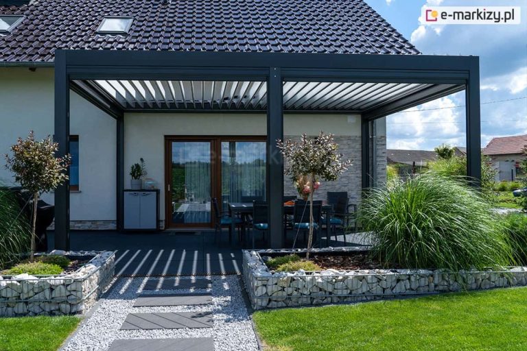 Nowoczesna, wolnostojąca pergola tarasowa Sb400 o konstrukcji aluminiowej w kolorze antracytowym, usytuowana przy domu z dachem krytym czarną dachówką i jasną elewacją. Dach pergoli składa się z ruchomych lameli bioklimatycznych w kolorze białym, które są częściowo otwarte, rzucając pasiaste cienie na ciemny taras. Pod zadaszeniem widoczny jest stół z krzesłami oraz fragment elewacji wyłożonej jasnym kamieniem dekoracyjnym. Na pierwszym planie znajdują się gabiony wypełnione jasnym kamieniem, z których wyrastają ozdobne trawy oraz dwa młode drzewka liściaste. Przez środek ogrodu prowadzi ścieżka z szarych, prostokątnych płyt ułożonych na jasnym żwirze, otoczona zielonym trawnikiem. W tle widoczne są fragmenty sąsiednich zabudowań pod błękitnym niebem z białymi chmurami.