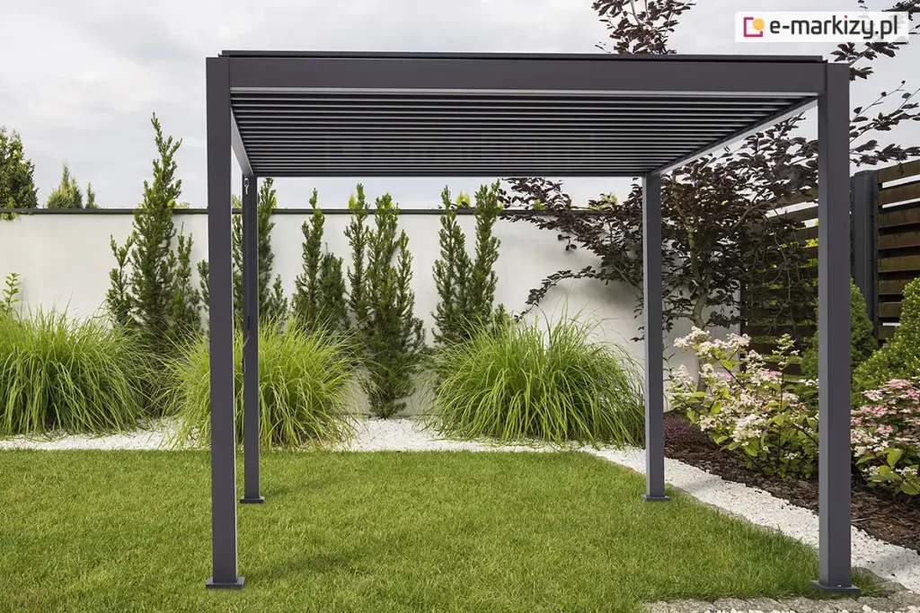 Wolnostojąca pergola tarasowa Gloria w kolorze ciemnego antracytu, ustawiona w estetycznie zaaranżowanym ogrodzie. Konstrukcja posiada cztery pionowe słupy o kwadratowym przekroju, które podtrzymują prostokątną ramę zadaszenia z widocznym systemem poziomych, ruchomych lameli. Lamele są ustawione w pozycji zamkniętej, tworząc jednolitą, płaską płaszczyznę dachu. Pergola jest zamontowana na trawniku oraz pasie jasnego grysu, który oddziela zieloną murawę od białego muru ogrodzeniowego. W tle widoczne są starannie dobrane rośliny ozdobne, w tym wysokie trawy, formowane iglaki oraz kwitnące krzewy hortensji. Całość kompozycji utrzymana jest w nowoczesnym, minimalistycznym stylu, a zachmurzone niebo zapewnia miękkie, równomierne oświetlenie konstrukcji.