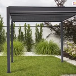 Wolnostojąca pergola tarasowa Gloria w kolorze ciemnego antracytu, ustawiona w estetycznie zaaranżowanym ogrodzie. Konstrukcja posiada cztery pionowe słupy o kwadratowym przekroju, które podtrzymują prostokątną ramę zadaszenia z widocznym systemem poziomych, ruchomych lameli. Lamele są ustawione w pozycji zamkniętej, tworząc jednolitą, płaską płaszczyznę dachu. Pergola jest zamontowana na trawniku oraz pasie jasnego grysu, który oddziela zieloną murawę od białego muru ogrodzeniowego. W tle widoczne są starannie dobrane rośliny ozdobne, w tym wysokie trawy, formowane iglaki oraz kwitnące krzewy hortensji. Całość kompozycji utrzymana jest w nowoczesnym, minimalistycznym stylu, a zachmurzone niebo zapewnia miękkie, równomierne oświetlenie konstrukcji.