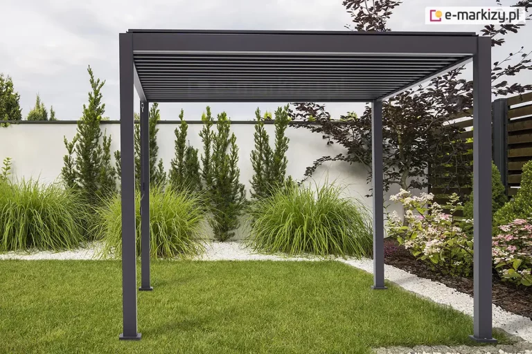 Wolnostojąca pergola tarasowa Gloria w kolorze ciemnego antracytu, ustawiona w estetycznie zaaranżowanym ogrodzie. Konstrukcja posiada cztery pionowe słupy o kwadratowym przekroju, które podtrzymują prostokątną ramę zadaszenia z widocznym systemem poziomych, ruchomych lameli. Lamele są ustawione w pozycji zamkniętej, tworząc jednolitą, płaską płaszczyznę dachu. Pergola jest zamontowana na trawniku oraz pasie jasnego grysu, który oddziela zieloną murawę od białego muru ogrodzeniowego. W tle widoczne są starannie dobrane rośliny ozdobne, w tym wysokie trawy, formowane iglaki oraz kwitnące krzewy hortensji. Całość kompozycji utrzymana jest w nowoczesnym, minimalistycznym stylu, a zachmurzone niebo zapewnia miękkie, równomierne oświetlenie konstrukcji.