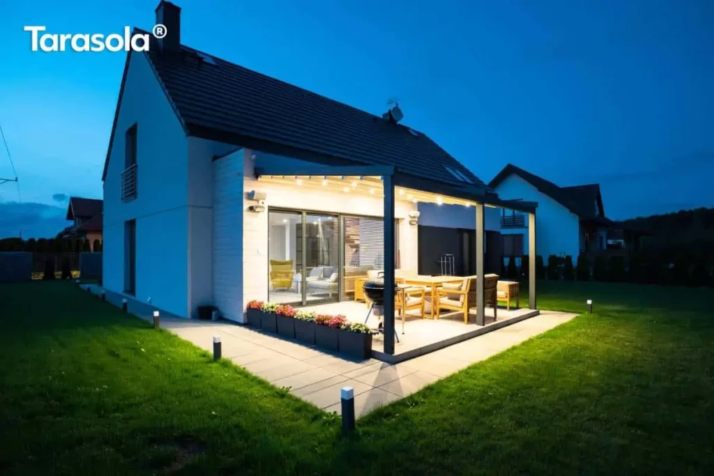 Nocna panorama nowoczesnego domu jednorodzinnego z zamontowaną przyścienną pergolą Elegancy Tarasola, która stanowi centralny punkt oświetlonego tarasu. Dwumodułowa konstrukcja w kolorze antracytowym posiada w pełni rozwinięte zadaszenie tkaninowe, pod którym świecą liczne zintegrowane punkty LED, oświetlając zestaw drewnianych mebli ogrodowych oraz czarny grill. Jasna elewacja budynku z drewnianymi akcentami kontrastuje z ciemnym, dwuspadowym dachem. Wokół tarasu rozmieszczono niskie donice z kwiatami oraz nowoczesne słupki oświetleniowe wyznaczające krawędź chodnika i trawnika. W tle, na rozległym ogrodzie, widoczne są zarysy sąsiednich domów pod nocnym, błękitnym niebem, co podkreśla spójność architektoniczną całego założenia.
