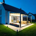 Nocna panorama nowoczesnego domu jednorodzinnego z zamontowaną przyścienną pergolą Elegancy Tarasola, która stanowi centralny punkt oświetlonego tarasu. Dwumodułowa konstrukcja w kolorze antracytowym posiada w pełni rozwinięte zadaszenie tkaninowe, pod którym świecą liczne zintegrowane punkty LED, oświetlając zestaw drewnianych mebli ogrodowych oraz czarny grill. Jasna elewacja budynku z drewnianymi akcentami kontrastuje z ciemnym, dwuspadowym dachem. Wokół tarasu rozmieszczono niskie donice z kwiatami oraz nowoczesne słupki oświetleniowe wyznaczające krawędź chodnika i trawnika. W tle, na rozległym ogrodzie, widoczne są zarysy sąsiednich domów pod nocnym, błękitnym niebem, co podkreśla spójność architektoniczną całego założenia.