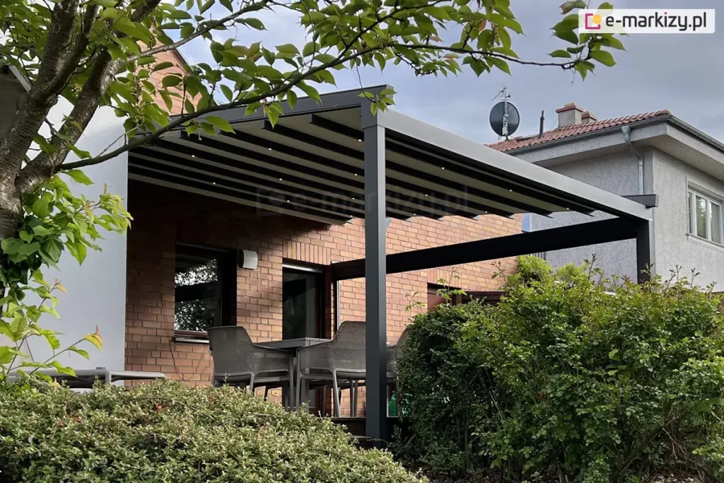 Nowoczesna pergola tarasowa Square o solidnej konstrukcji aluminiowej w kolorze ciemnego antracytu, zamontowana do elewacji budynku wykonanej z czerwonej cegły klinkierowej. Zadaszenie pergoli to rozsuwana tkanina w kolorze białym lub jasnoszarym, która po rozwinięciu układa się w płaskie, skośne zadaszenie, a pod jej profilami poprzecznymi zintegrowano punktowe oświetlenie LED. Konstrukcja wspiera się na grubym, pionowym słupie nośnym, a boczne ramy tworzą prostokątne obramowanie przestrzeni wypoczynkowej. Na tarasie pod zadaszeniem widoczny jest zestaw mebli ogrodowych, w tym nowoczesne krzesła o ażurowej strukturze w kolorze szarym, ustawione wokół stołu. Teren wokół tarasu jest gęsto porośnięty naturalną roślinnością, w tym zielonymi krzewami oraz drzewem liściastym na pierwszym planie, którego gałęzie częściowo przesłaniają widok na konstrukcję. W tle widoczny jest sąsiedni budynek mieszkalny o jasnej elewacji z dachówką oraz zamontowana na dachu antena satelitarna. Zdjęcie wykonane przy lekko zachmurzonym niebie, prezentujące funkcjonalne wykorzystanie pergoli jako osłony nad zewnętrzną strefą jadalnianą.