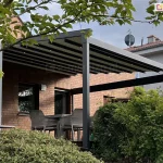 Nowoczesna pergola tarasowa Square o solidnej konstrukcji aluminiowej w kolorze ciemnego antracytu, zamontowana do elewacji budynku wykonanej z czerwonej cegły klinkierowej. Zadaszenie pergoli to rozsuwana tkanina w kolorze białym lub jasnoszarym, która po rozwinięciu układa się w płaskie, skośne zadaszenie, a pod jej profilami poprzecznymi zintegrowano punktowe oświetlenie LED. Konstrukcja wspiera się na grubym, pionowym słupie nośnym, a boczne ramy tworzą prostokątne obramowanie przestrzeni wypoczynkowej. Na tarasie pod zadaszeniem widoczny jest zestaw mebli ogrodowych, w tym nowoczesne krzesła o ażurowej strukturze w kolorze szarym, ustawione wokół stołu. Teren wokół tarasu jest gęsto porośnięty naturalną roślinnością, w tym zielonymi krzewami oraz drzewem liściastym na pierwszym planie, którego gałęzie częściowo przesłaniają widok na konstrukcję. W tle widoczny jest sąsiedni budynek mieszkalny o jasnej elewacji z dachówką oraz zamontowana na dachu antena satelitarna. Zdjęcie wykonane przy lekko zachmurzonym niebie, prezentujące funkcjonalne wykorzystanie pergoli jako osłony nad zewnętrzną strefą jadalnianą.