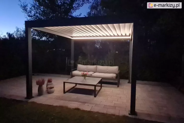 Zdjęcie przedstawia wolnostojącą pergolę bioklimatyczną o nowoczesnej, minimalistycznej konstrukcji, sfotografowaną w porze wieczornej. Rama pergoli oraz cztery masywne słupy nośne o kwadratowym przekroju wykonane są z aluminium w kolorze głębokiego, matowego antracytu. Zadaszenie składa się z równolegle ułożonych, białych lameli, które są podświetlone od spodu zintegrowanym systemem oświetlenia LED w postaci liniowych taśm zamontowanych wzdłuż wewnętrznej ramy. Światło o ciepłej barwie intensywnie rozświetla wnętrze pergoli, kontrastując z zapadającym zmrokiem i ciemną ścianą drzew w tle. Pod konstrukcją, na podłożu z jasnej kostki brukowej o nieregularnych odcieniach beżu, znajduje się strefa wypoczynkowa wyposażona w długą sofę z jasnoszarymi poduszkami oraz niski stolik kawowy z ciemnym stelażem i blatem w kolorze szarym. Po lewej stronie, obok mebli, ustawiono dekoracyjne donice z suchą roślinnością. Całość aranżacji tworzy luksusową i nastrojową przestrzeń ogrodową, idealną do relaksu po zachodzie słońca.