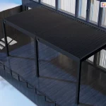 Dwumodułowa pergola lamelowa Marlo w kolorze antracytowym, zamontowana do elewacji nowoczesnego budynku z dużymi przeszkleniami. Konstrukcja składa się z dwóch połączonych sekcji zadaszenia, które wspierają się na trzech pionowych słupach o kwadratowym przekroju, ustawionych w jednej linii od frontu. Lamele dachowe są całkowicie zamknięte, tworząc jednolitą, ciemną płaszczyznę chroniącą rozległy taras. Taras został wykonany z ciemnych desek o wyraźnym połysku, a jego krawędź zabezpieczono minimalistyczną, ciemną balustradą. Po lewej stronie widoczne są schody prowadzące na niższy poziom oraz dekoracyjne, bezlistne drzewko w białym kolorze, co sugeruje zimową lub wczesnowiosenną scenerię z zalegającym gdzieniegdzie śniegiem. Całość ujęcia wykonana jest z perspektywy górnej (z lotu ptaka), co pozwala dokładnie ocenić skalę podwójnego zadaszenia oraz jego integrację z bryłą budynku i otoczeniem.