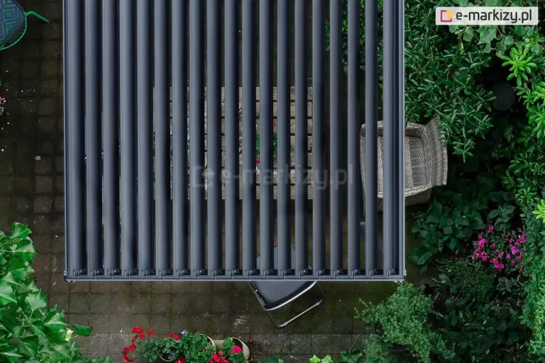 Widok prosto z góry (z lotu ptaka) na dach bioklimatycznej pergoli Gloria w kolorze antracytowym z otwartymi lamelami. Równolegle ustawione, wąskie panele aluminiowe tworzą rytmiczny wzór, przez który widać fragmenty wyposażenia tarasu: drewniany stół z zastawą oraz pleciony fotel ogrodowy. Pergola jest usytuowana na podłożu z ciemnej kostki brukowej, otoczona gęstą roślinnością ogrodową, krzewami o soczyście zielonych liściach oraz donicami z kwitnącymi na czerwono i różowo kwiatami. Perspektywa podkreśla funkcję regulacji dopływu światła oraz integrację nowoczesnej konstrukcji z naturalnym otoczeniem ogrodu.