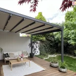 Zdjęcie prezentuje nowoczesną pergolę tarasową aura o konstrukcji aluminiowej w kolorze antracytowym, zamontowaną do jasnej elewacji budynku mieszkalnego. Pergola posiada rozsuwane zadaszenie tkaninowe w kolorze kremowym, które jest rozpięte na systemie poprzecznych profili jezdnych. W dolnej części profili zamontowano zintegrowane, punktowe oświetlenie LED, emitujące ciepłe światło. Konstrukcja wspiera się na pionowych słupach o kwadratowym przekroju, usytuowanych na rogu drewnianego tarasu. Pod pergolą zaaranżowano strefę wypoczynkową z meblami ogrodowymi o drewnianej strukturze i jasnych poduszkach, składającą się z sofy i fotela. Na meblach leżą poduszki dekoracyjne, w tym jedna czarna z białym napisem oraz jedna beżowa z etnicznym wzorem. Na tarasie z ciemnobrązowych desek leży jasny dywan z frędzlami, na którym stoi prostokątny, drewniany stolik kawowy z wazonem z różowymi kwiatami i dekoracyjnymi świecznikami. Po lewej stronie, przy ścianie budynku, zamocowana jest długa, czarna korba do manualnego sterowania zadaszeniem. W narożniku tarasu rośnie ozdobne drzewko o bordowych liściach w ciemnej donicy, a przy krawędzi ustawiono dwie okrągłe, betonowe donice z zielonymi trawami. Tło stanowi gęsty, zielony ogród z krzewami i drzewami liściastymi, a u góry kadru widoczne są gałązki z czerwonymi liśćmi, prawdopodobnie klonu palmowego, obramowujące ujęcie.