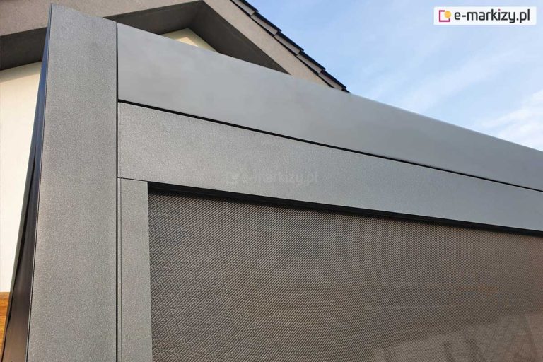 Zdjęcie przedstawia detal konstrukcyjny nowoczesnej pergoli aluminiowej w kolorze antracytowym o matowym, ziarnistym wykończeniu. Widoczne jest precyzyjne łączenie pionowego słupa wsporczego z poziomą belką ramy. Wewnątrz ramy zamontowany został refleksol Zip, którego tkanina o gęstym splocie w odcieniach brązu i szarości jest częściowo rozwinięta. Tkanina rolety porusza się w prowadnicach bocznych zintegrowanych z profilem słupa, co zapewnia stabilność materiału. W górnej części kadru widoczny jest fragment jasnej elewacji budynku oraz krawędź dachu, co obrazuje sposób montażu konstrukcji w bliskim sąsiedztwie obiektu. Zastosowane profile charakteryzują się minimalistycznym, prostokątnym przekrojem i wysoką estetyką wykonania detali.